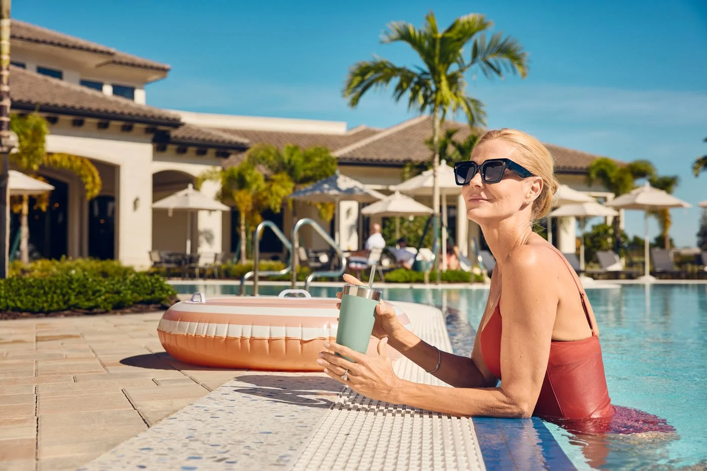 A woman in sunglasses and a red swimsuit rests at the pool edge holding a tumbler, a pink float beside her and the clubhouse behind.