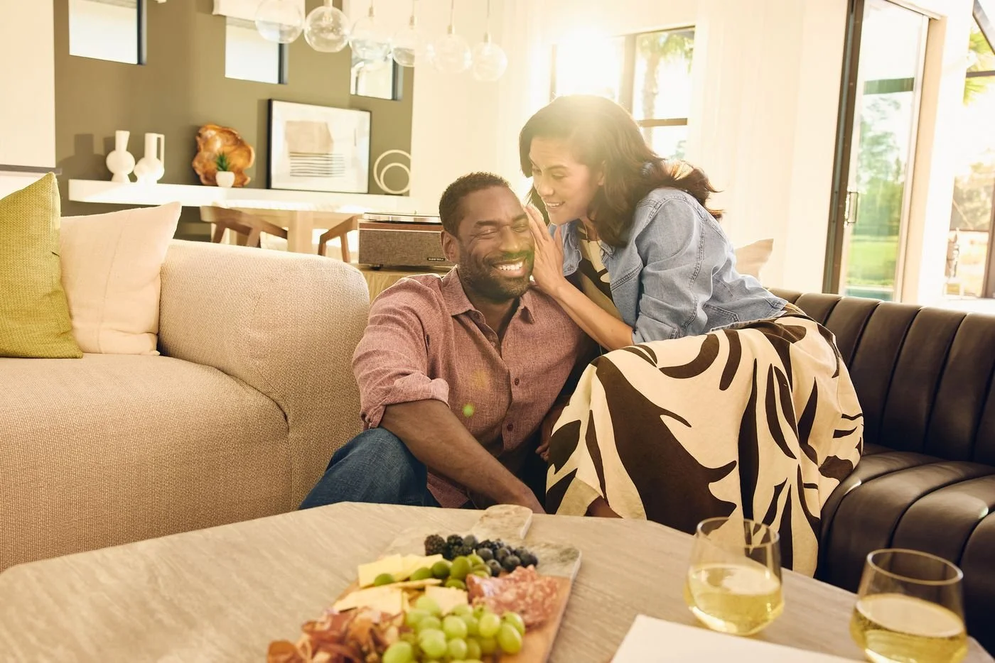 A woman leans in to whisper to a laughing man on a living room couch, a snack board on the table.