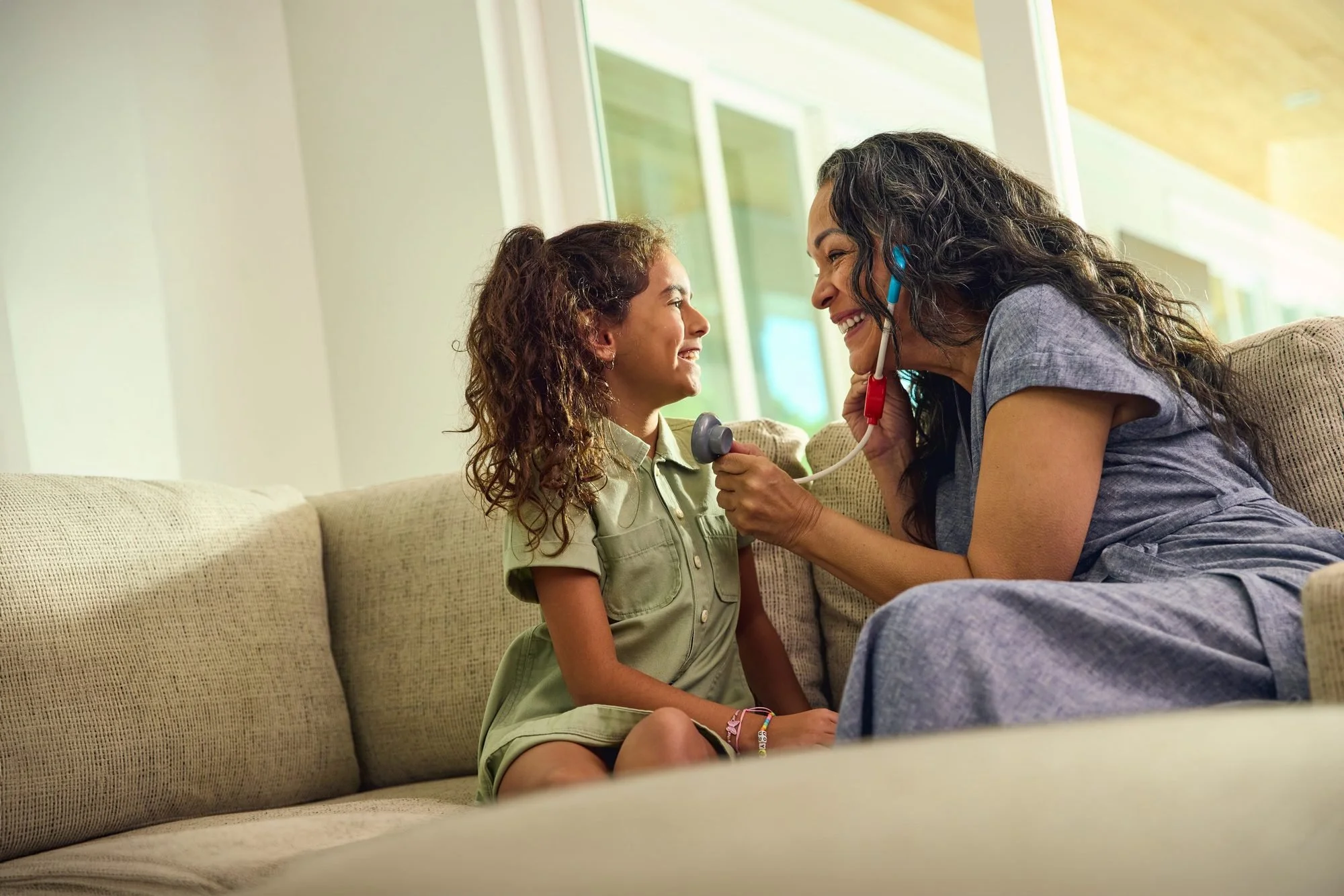 A girl and a woman laugh as they play doctor with a toy stethoscope on a couch.
