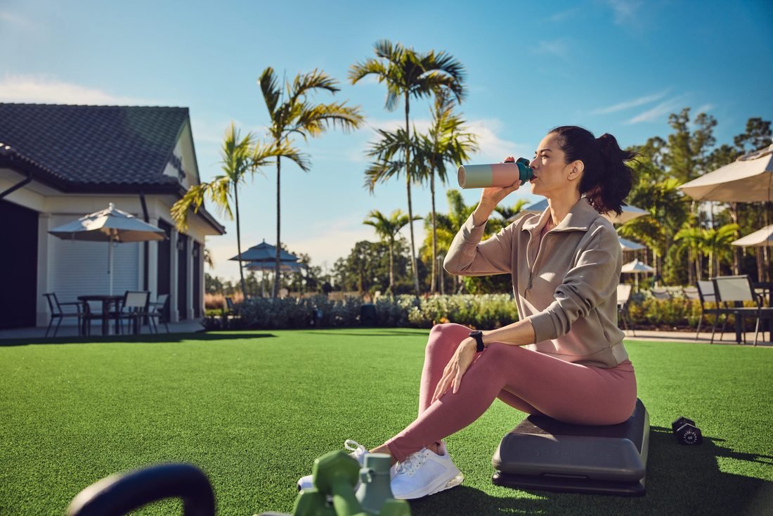 A woman in a half-zip pullover and leggings sits on a step platform and drinks from a shaker bottle on a turf lawn, a clubhouse behind her.