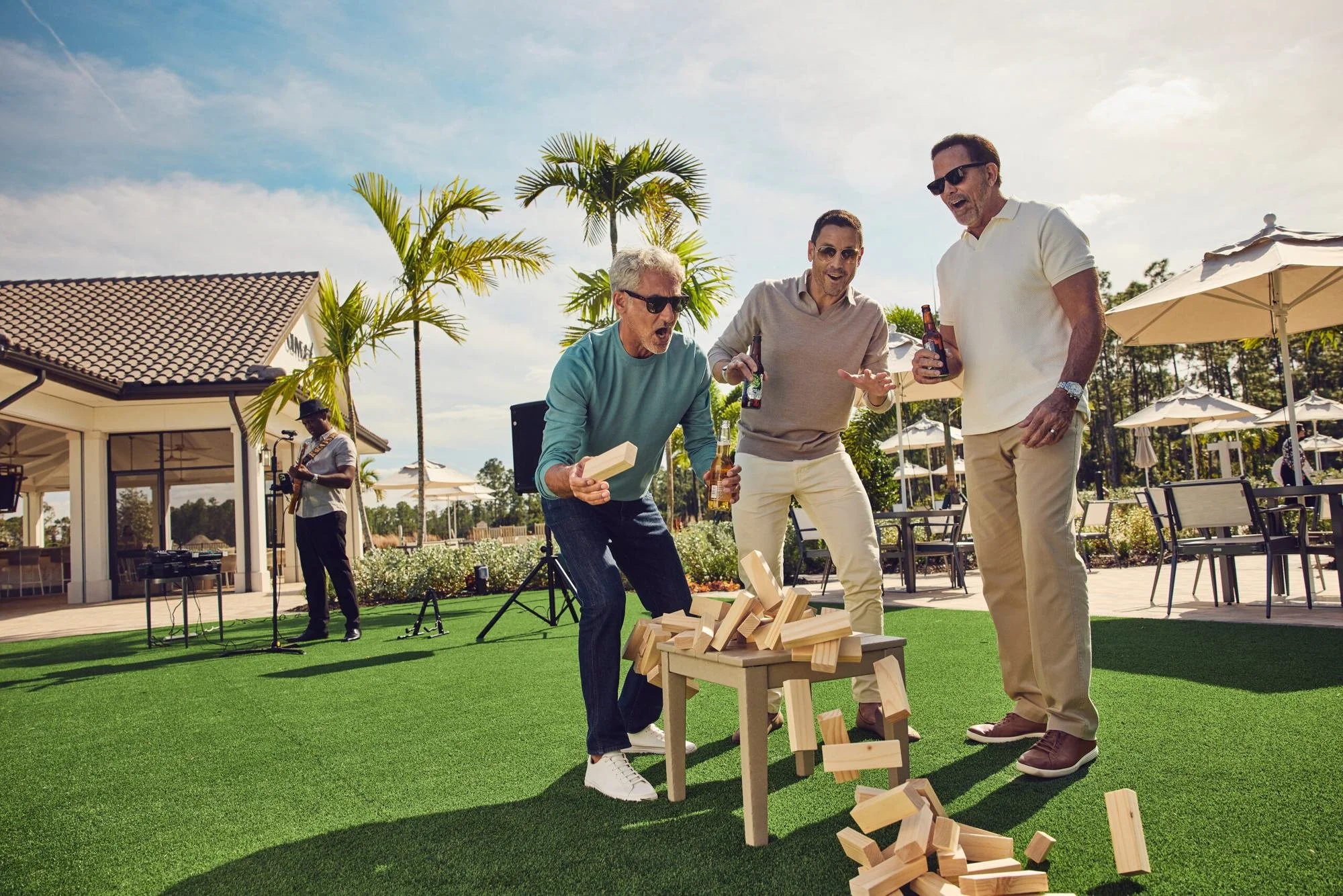 Three men laugh as a giant block tower falls on a clubhouse lawn.