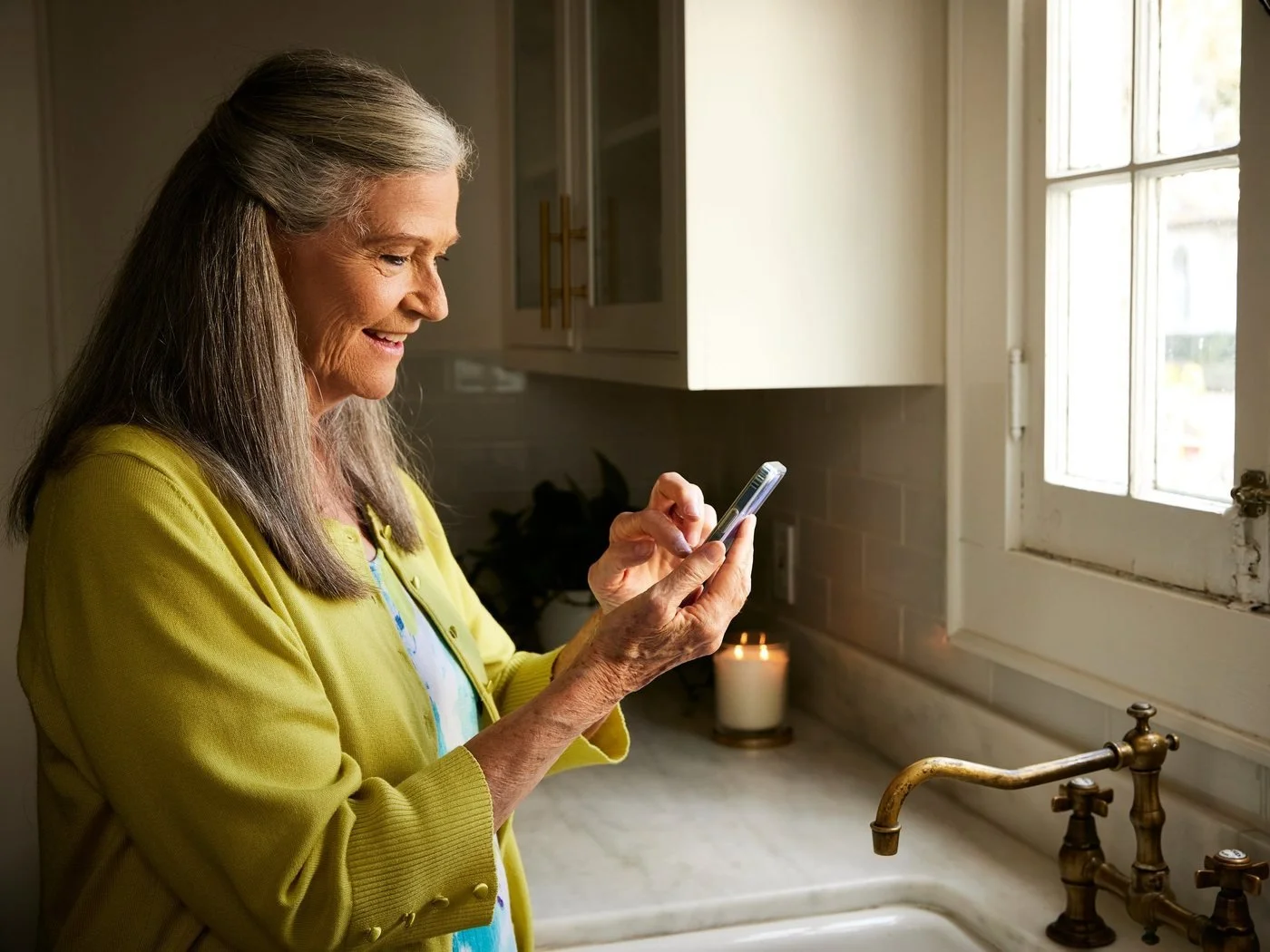A woman smiles at her phone while standing at a kitchen sink.