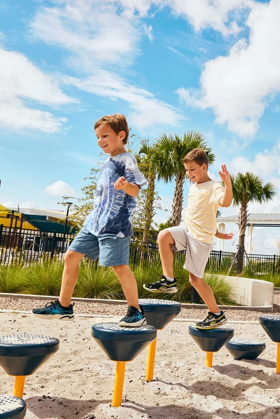 Two boys hop across stepping posts on a sandy playground.