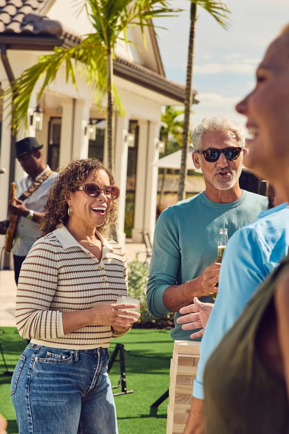 A woman with curly hair laughs with a drink beside a gray-haired man in sunglasses at an outdoor gathering, a guitarist behind them.
