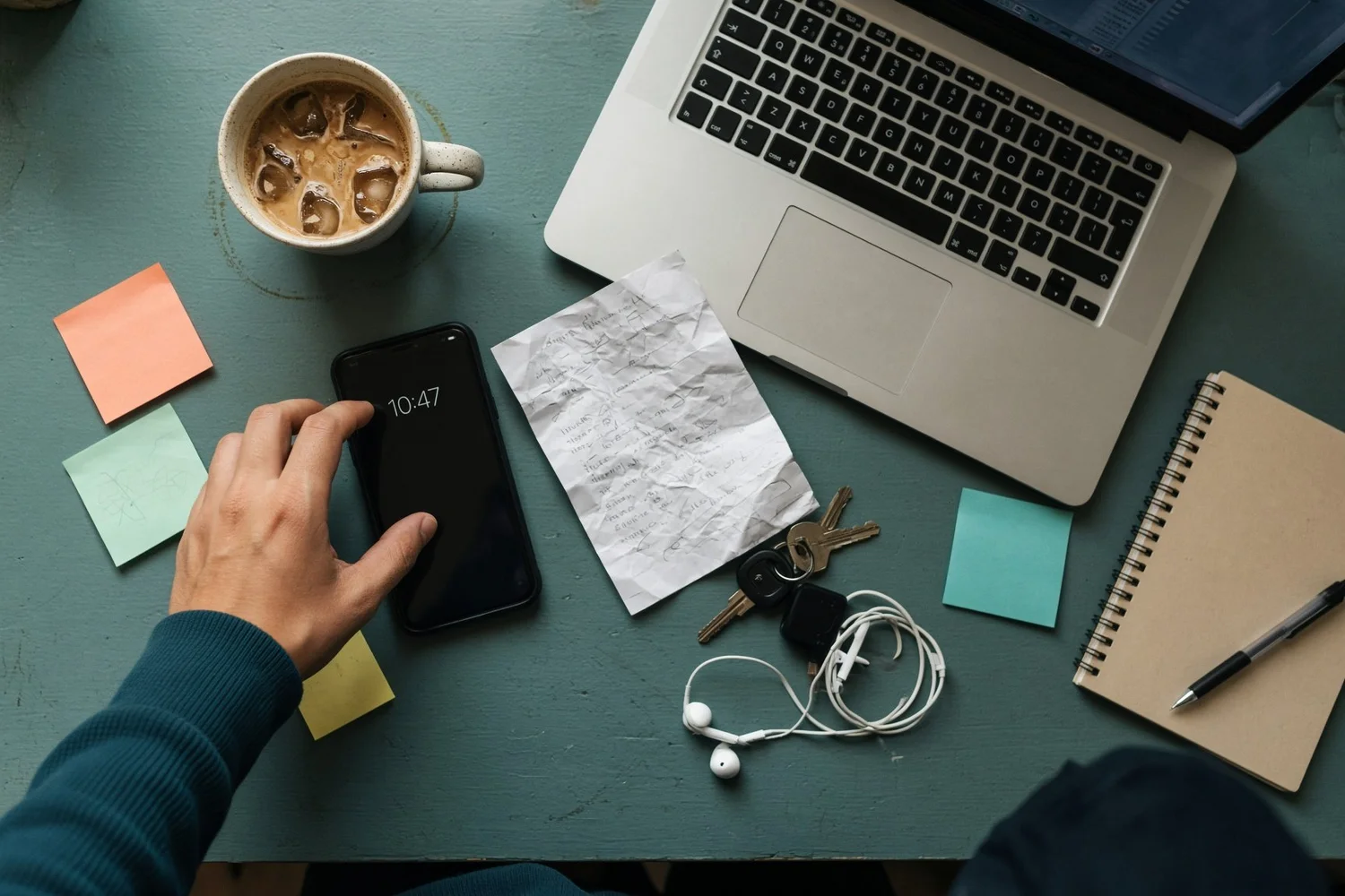 A cluttered desk with sticky notes, a cold coffee and a hand reaching for a phone showing the time