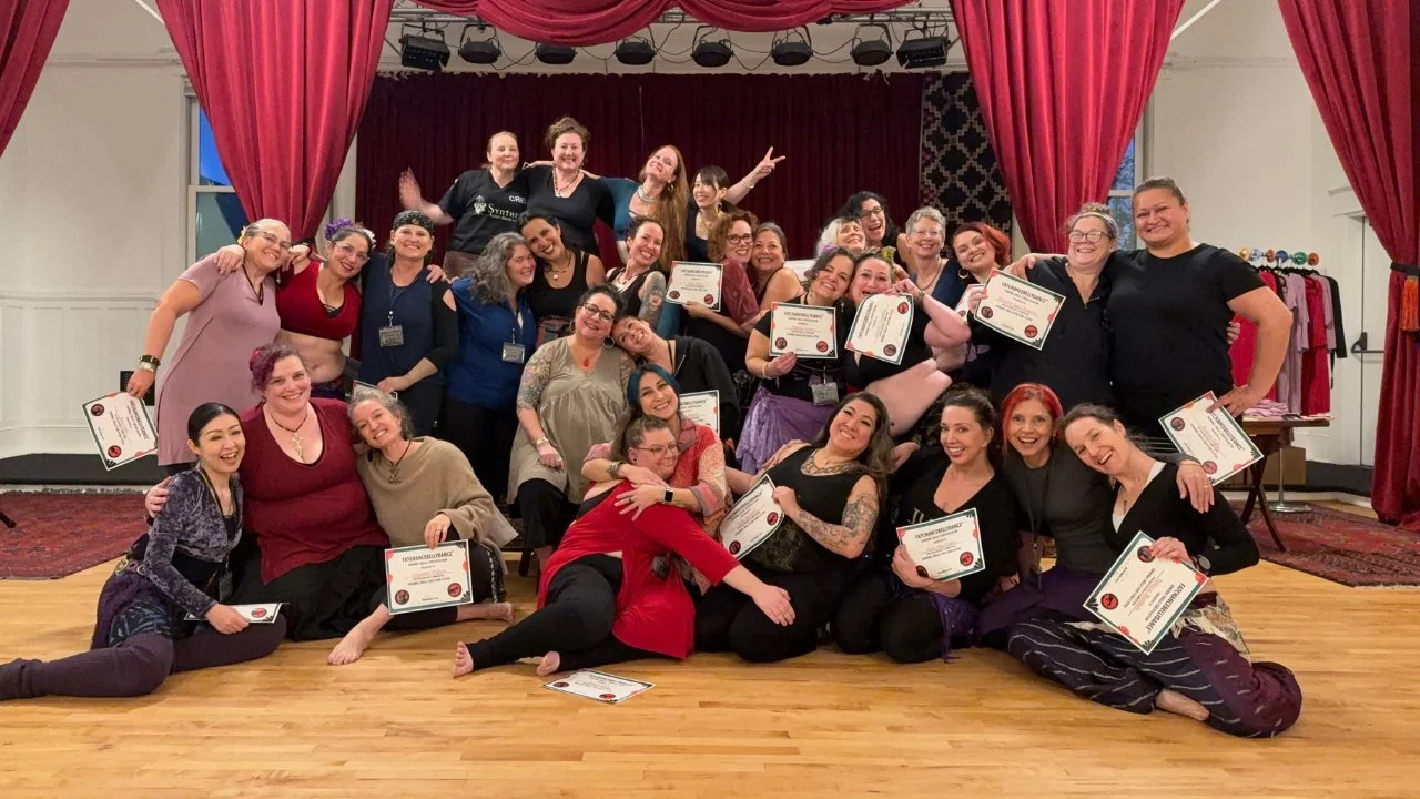 A large group of smiling General Skills graduates holding their FatChanceBellyDance® certificates on a studio stage