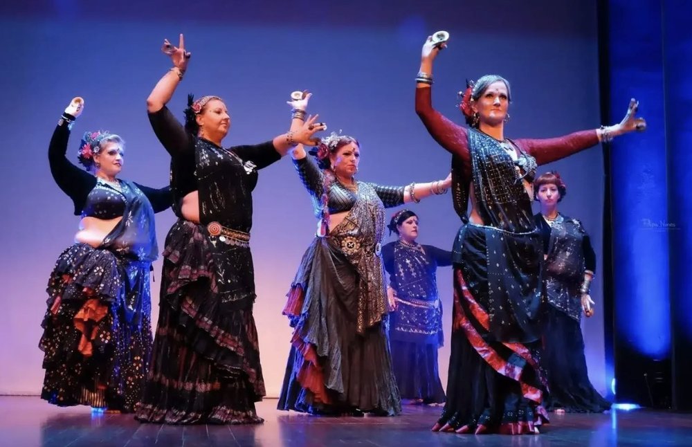 A troupe of FCBD® Style dancers performing together on stage, arms raised with zills