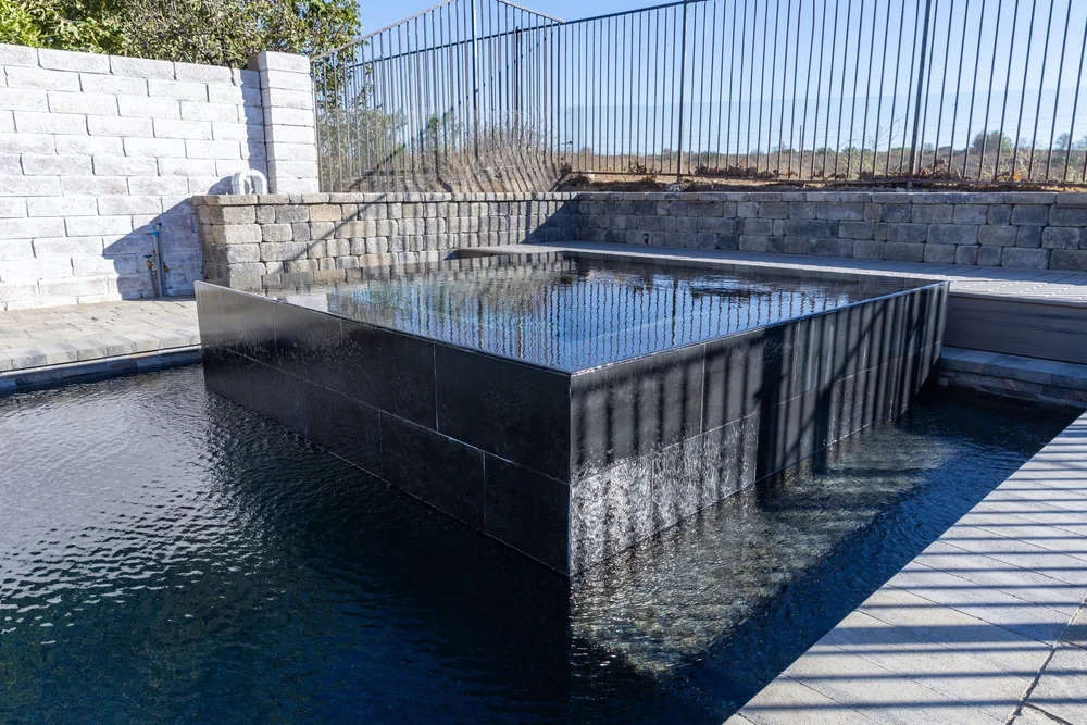 Raised square spa with dark tile sides beside the pool and gray paver deck.