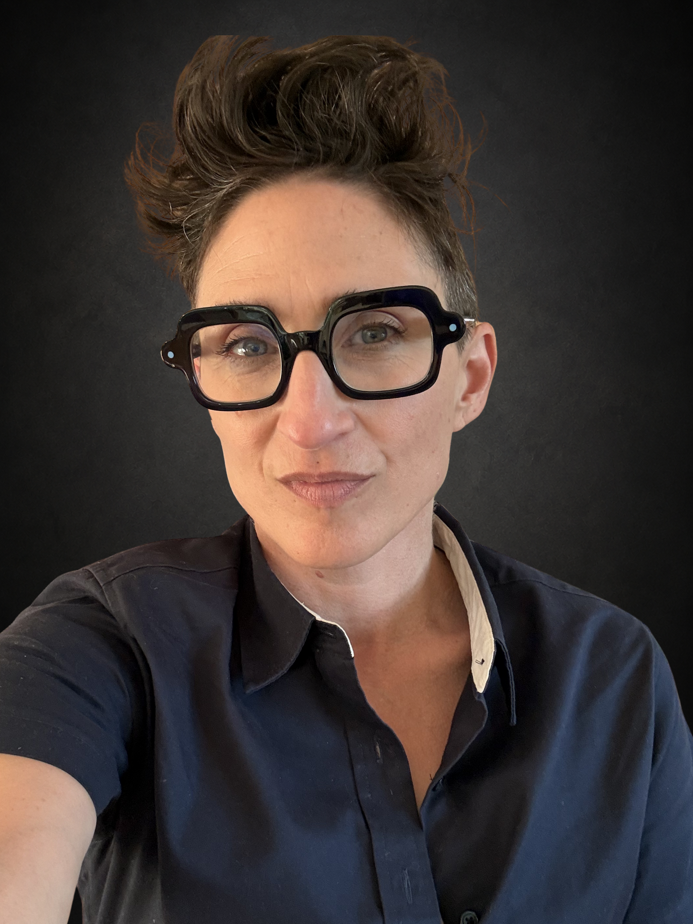 Tam looks at the camera with a slight smile, wearing black glasses and a dark button-up shirt against a dark charcoal background.