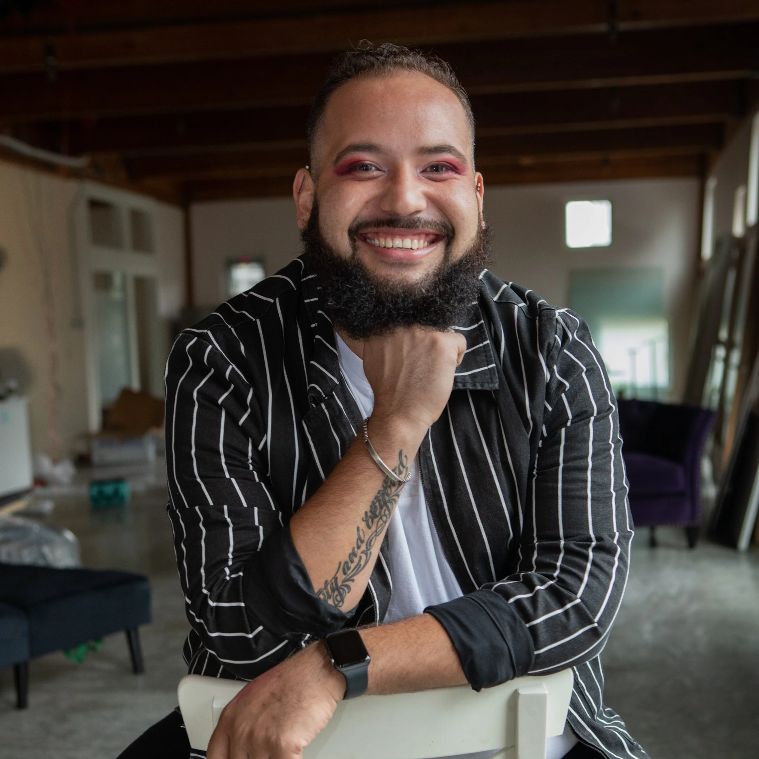 Malik smiles at the camera while seated on a chair, wearing a black striped shirt, with exposed wood beams and a softly blurred indoor space behind him.