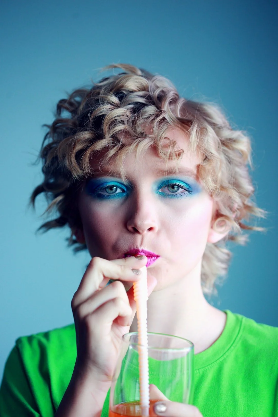 A person with curly blond hair, bright blue eye makeup, and a green shirt drinks from a striped straw.