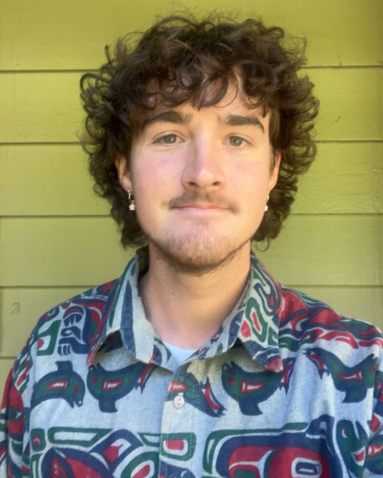 Jenner looks at the camera with a neutral expression, wearing earrings and a patterned button-up shirt in front of a green wood-paneled wall.