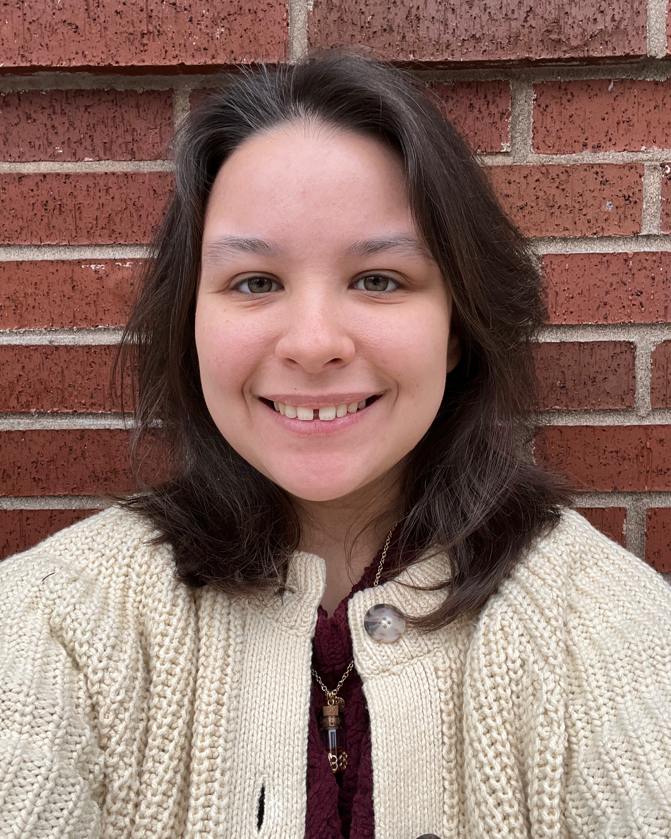 Ollie smiles at the camera in front of a red brick wall, wearing a cream knit cardigan and a pendant necklace.