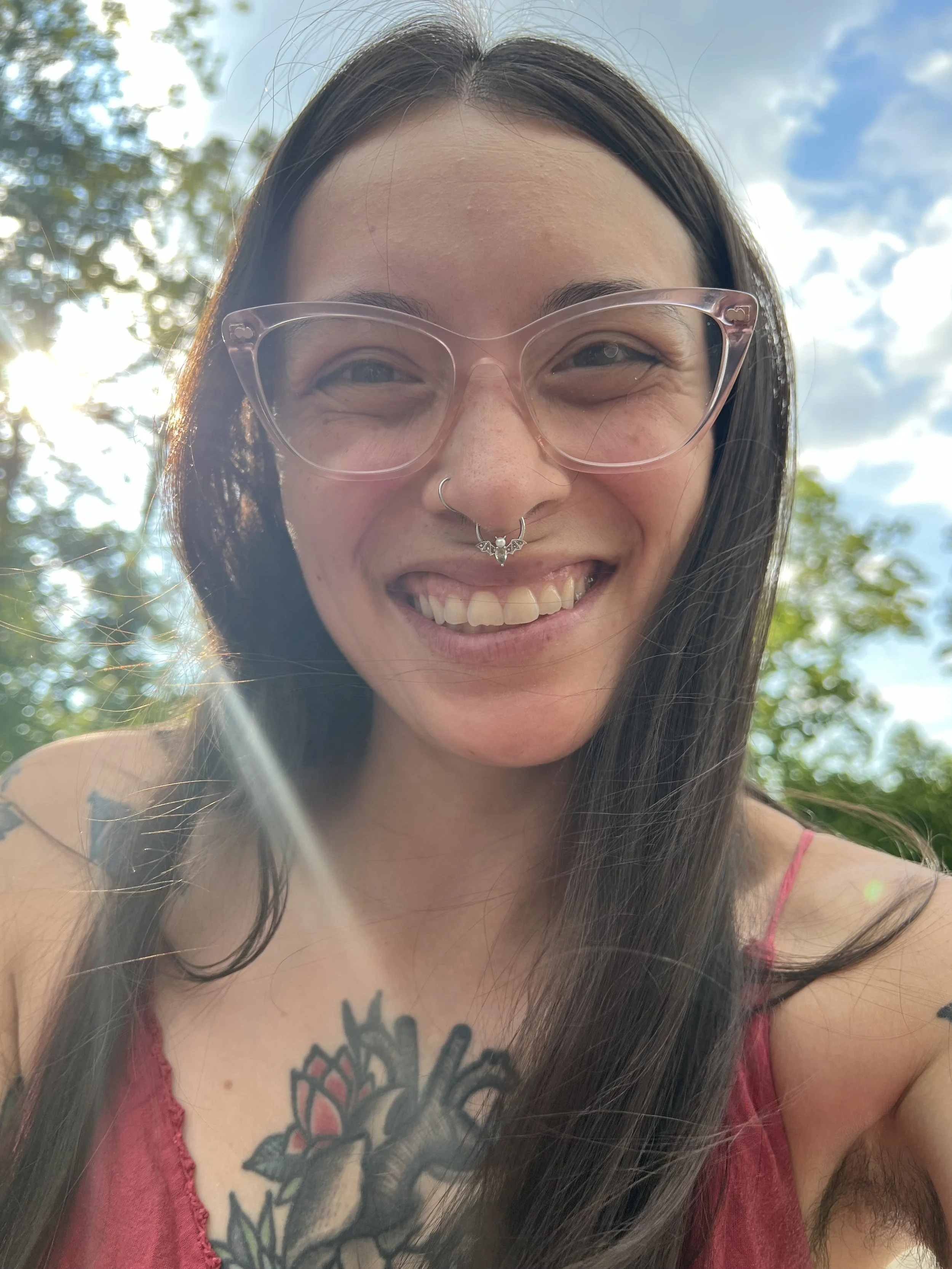 Aria smiles at the camera outdoors, wearing pink glasses and a septum ring, with trees and a bright sky in the background.