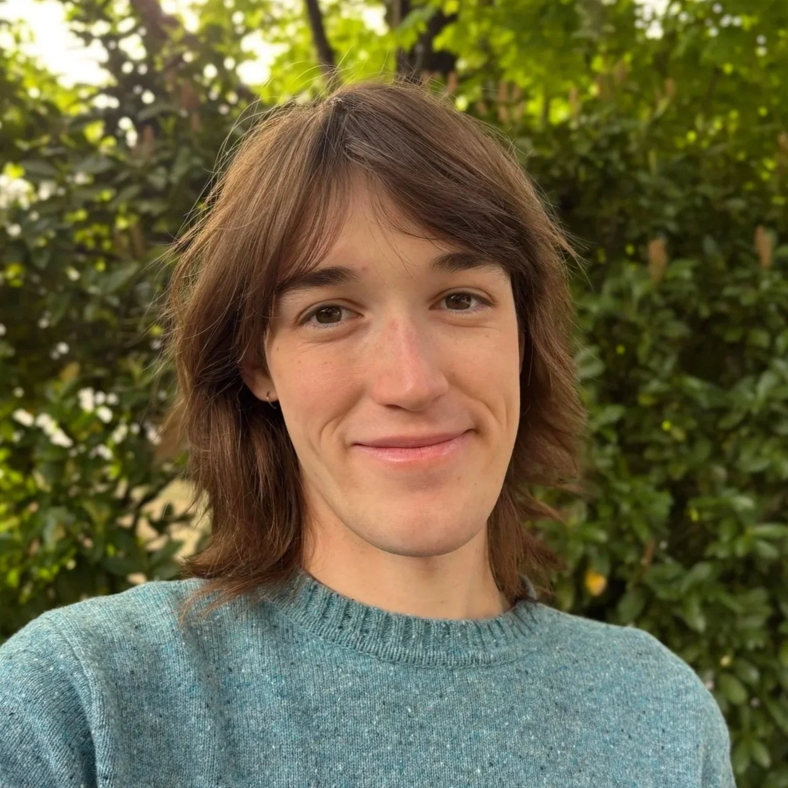 Taylor smiles softly at the camera outdoors, wearing a teal sweater with green trees and foliage in the background.