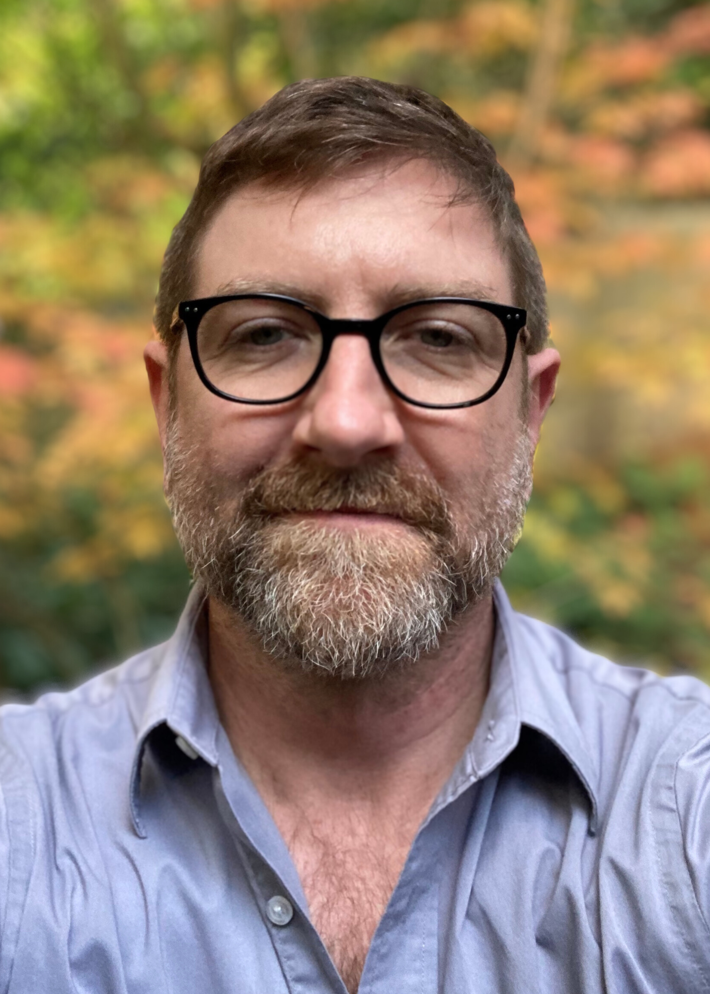 Matt looks at the camera with a slight smile, wearing black glasses and a light blue button-up shirt, with softly blurred trees in the background.