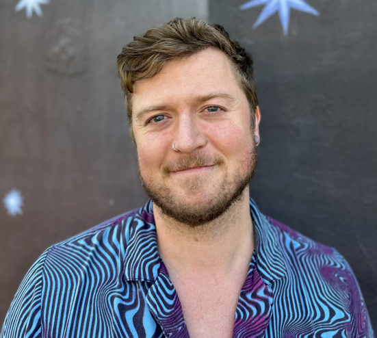 Chase looks at the camera with a slight smile, wearing a blue and purple patterned shirt in front of a dark wall with small painted stars.