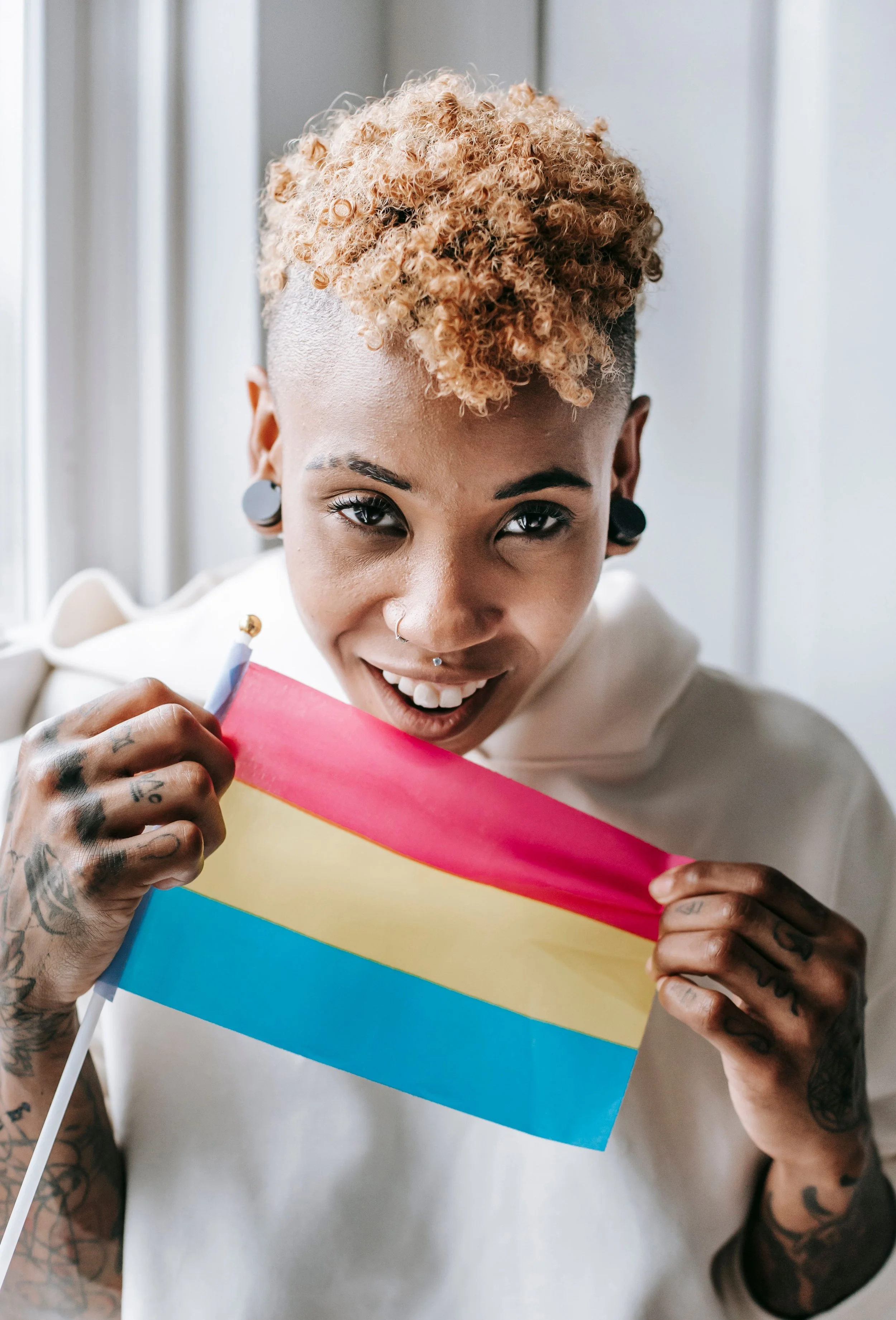A transmasc Black person smiles while holding a Pansexual Pride flag around their shoulders. Credit: Photo by Tim Samuel from Pexels.