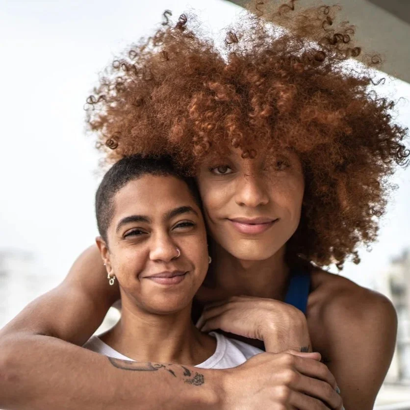 Two people stand close together, smiling softly at the camera, with one person’s arms wrapped around the other. The background is bright and softly blurred.