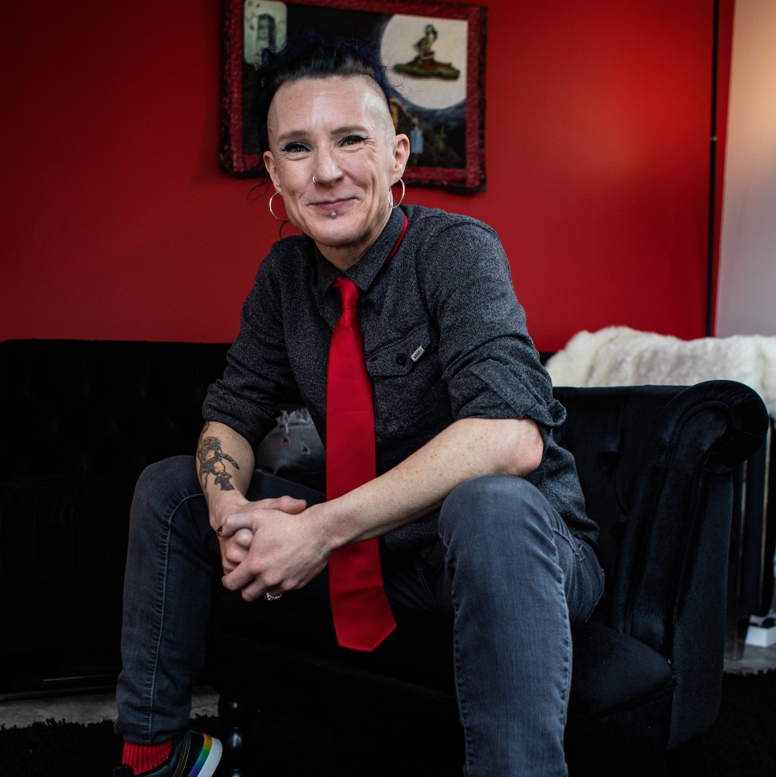 Morgan smiles at the camera while seated on a black couch, wearing a gray button-up shirt and bright red tie against a red wall.