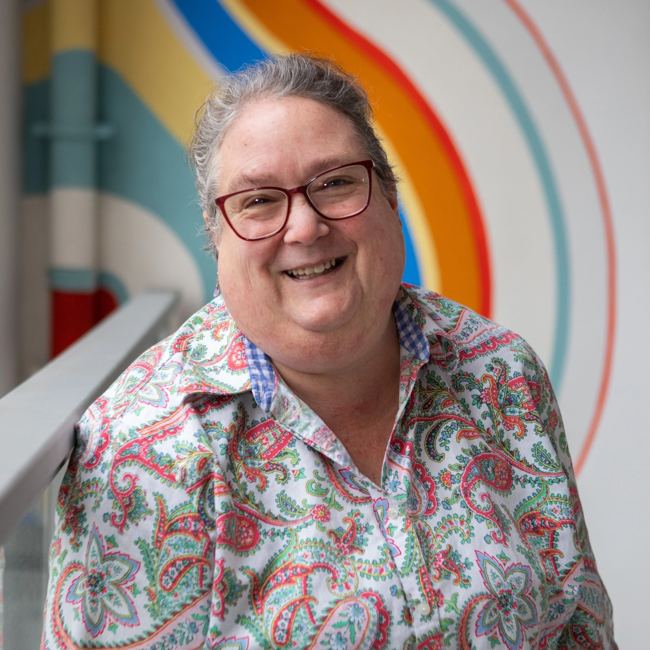 Irene smiles at the camera, wearing red glasses and a colorful paisley shirt, with a bright rainbow mural in the background.