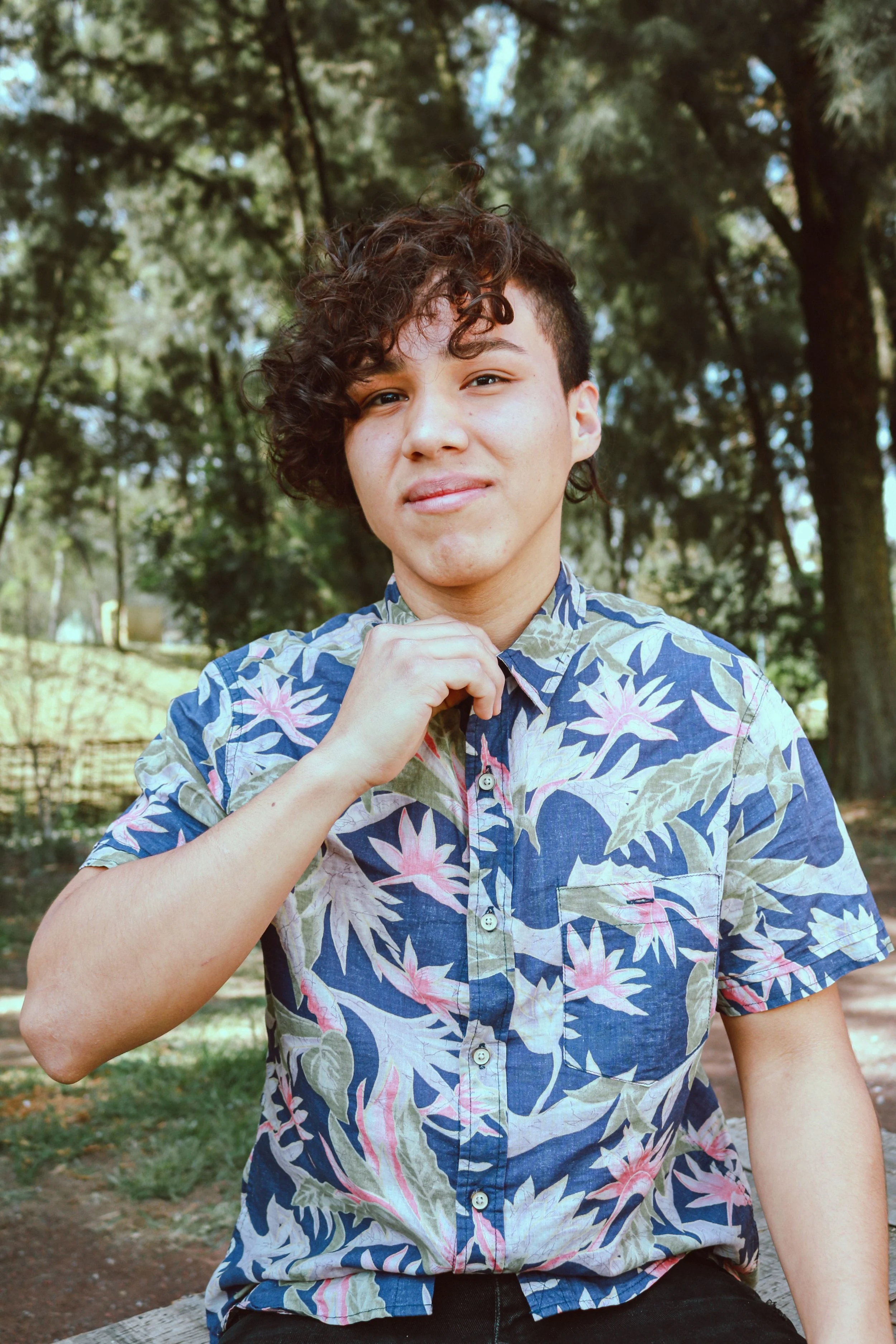 A trans masc person adjusts their shirt collar while standing outdoors in a sunny park. Credit: Photo by Jonathan Valdes from Pexels.