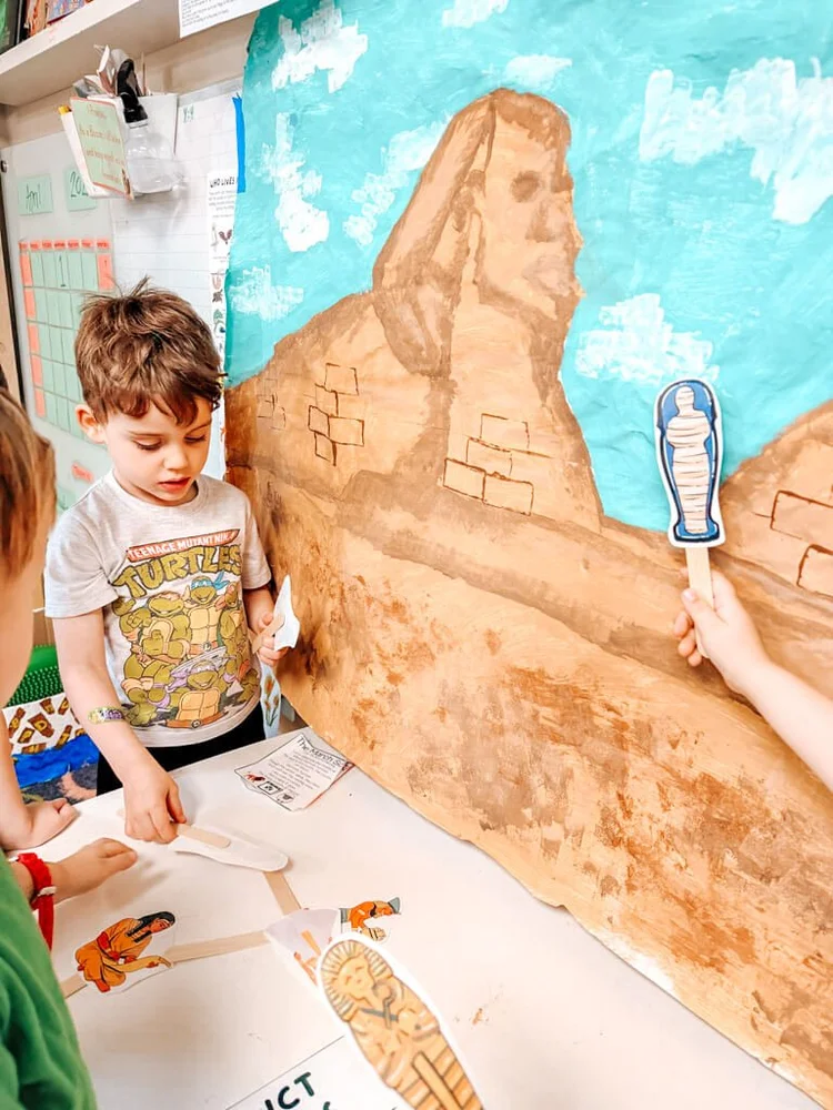 Preschoolers using stick puppets to learn about ancient Egypt in front of a painted Sphinx mural