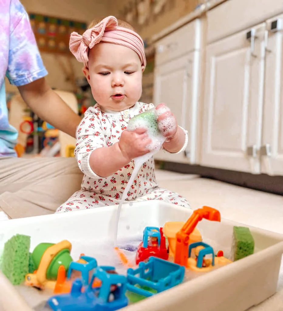 Baby smiling at a soapy sensory bin with toy trucks