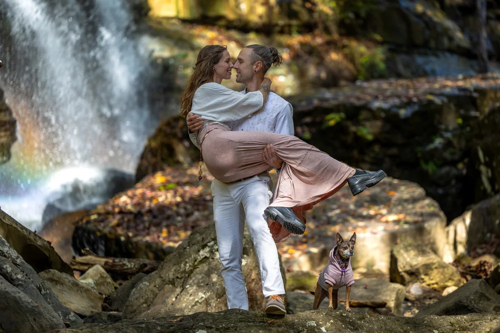 Couple embracing beside a waterfall with a rainbow, their dog watching