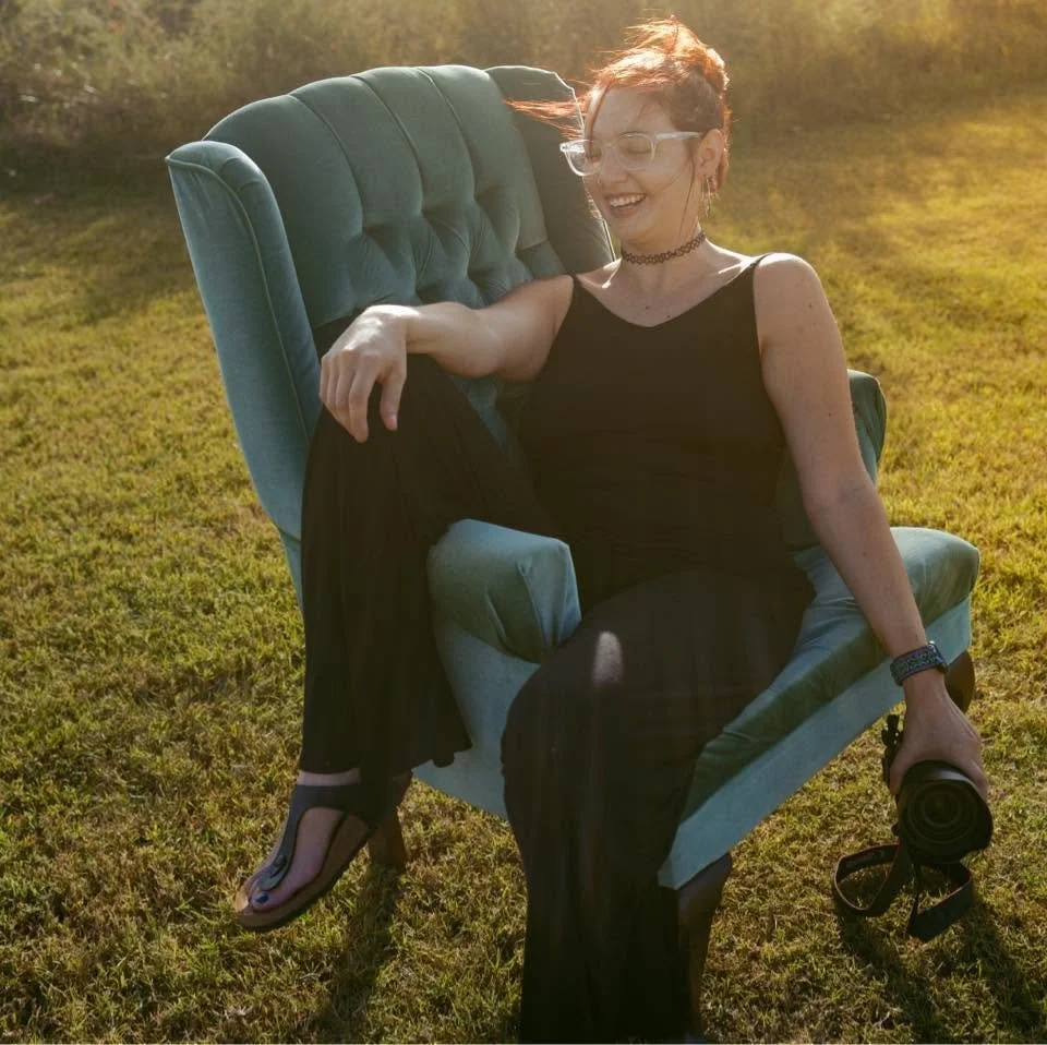 Zoe laughing in a velvet armchair in a sunlit field, camera in hand
