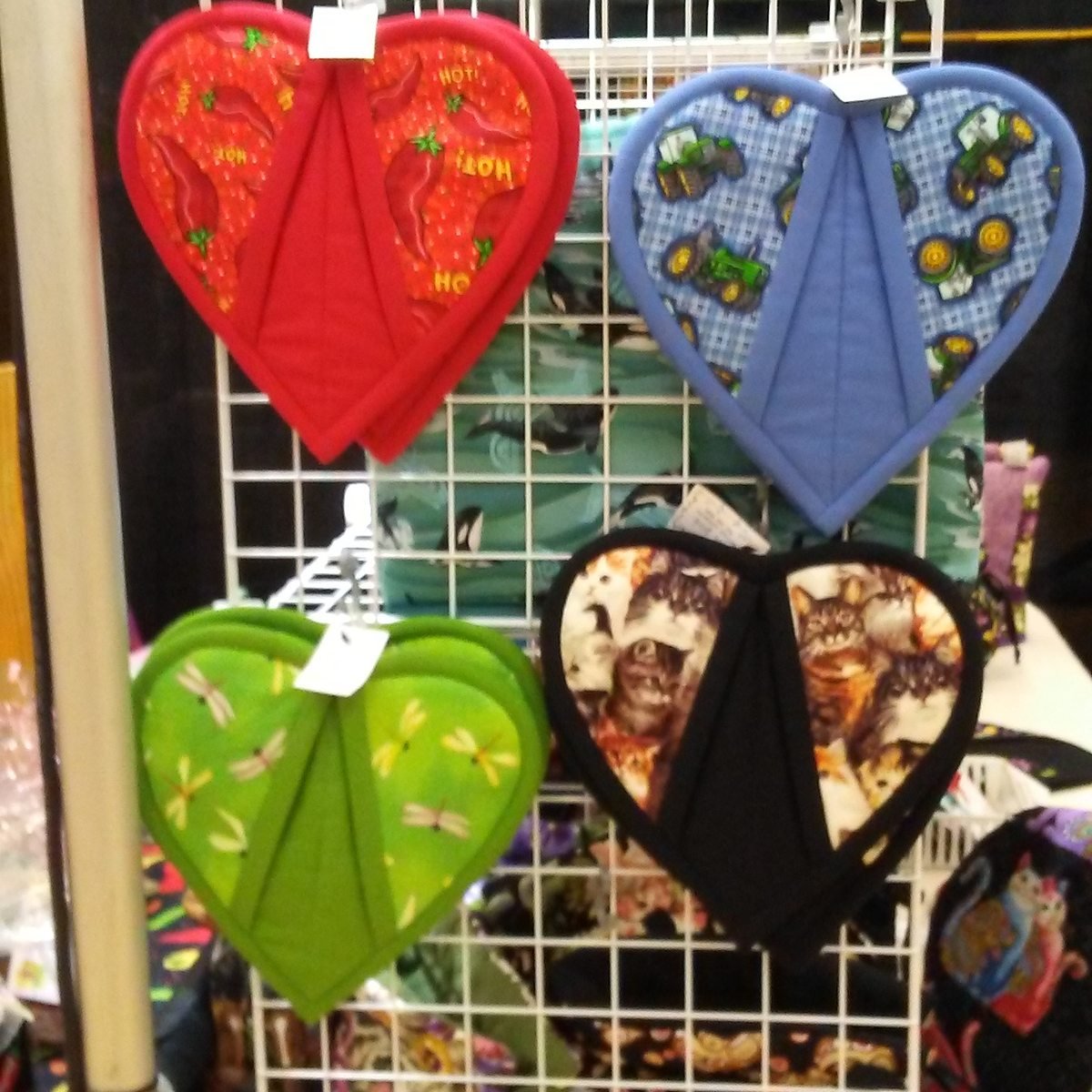 Four heart-shaped quilted pot holders hanging on a wire rack, in red chilli, blue tractor, green dragonfly and black cat prints