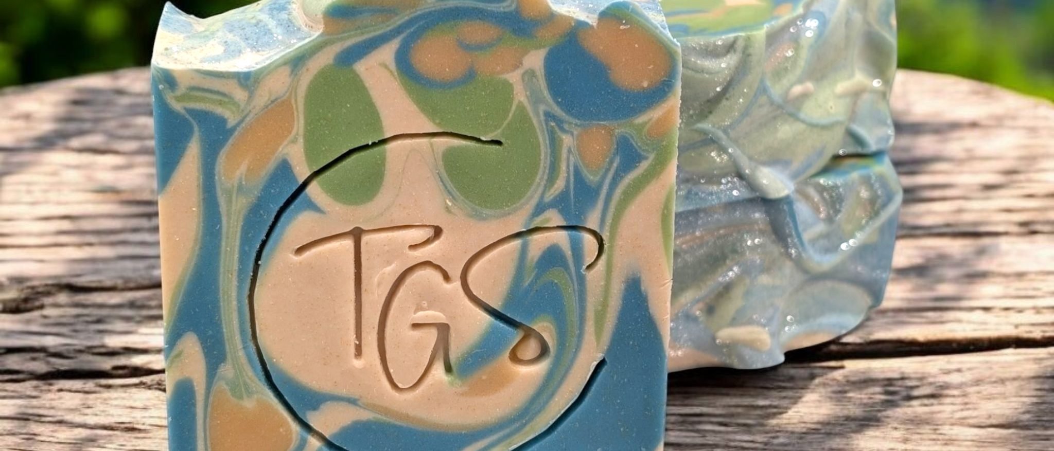 A bar of handmade soap swirled in blue, green and cream, stamped with the Triple Goddess Soapery mark, resting on weathered wood