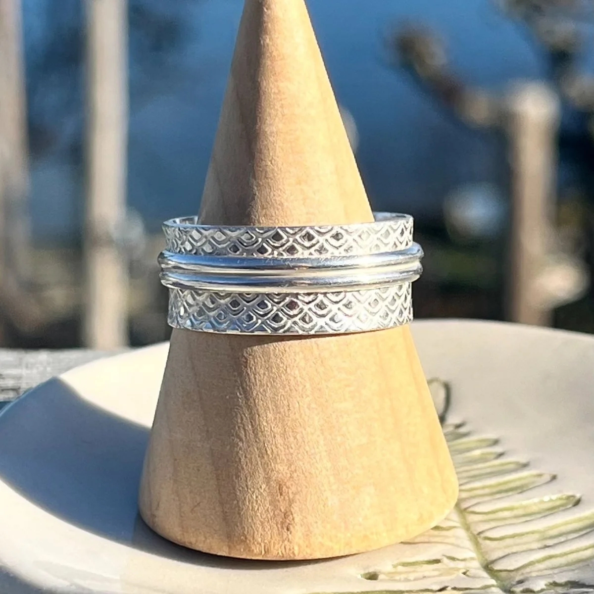A wide sterling silver band stamped with a diamond lattice pattern, on a wooden ring cone resting on a handmade ceramic dish