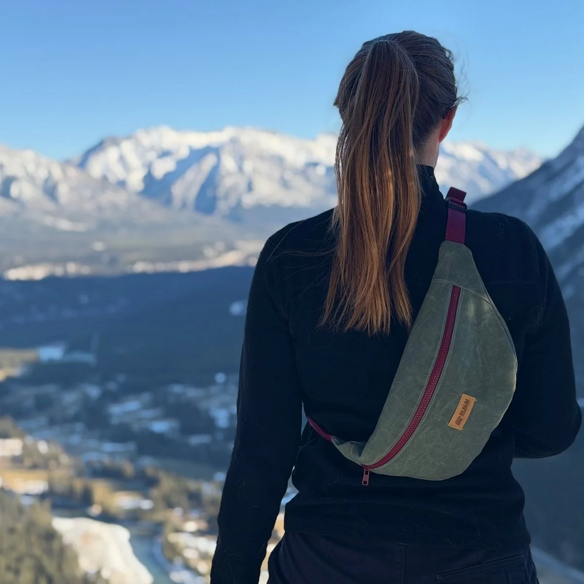 A woman seen from behind wearing a sage waxed canvas sling bag with a wine red zip, looking out over a snowy valley
