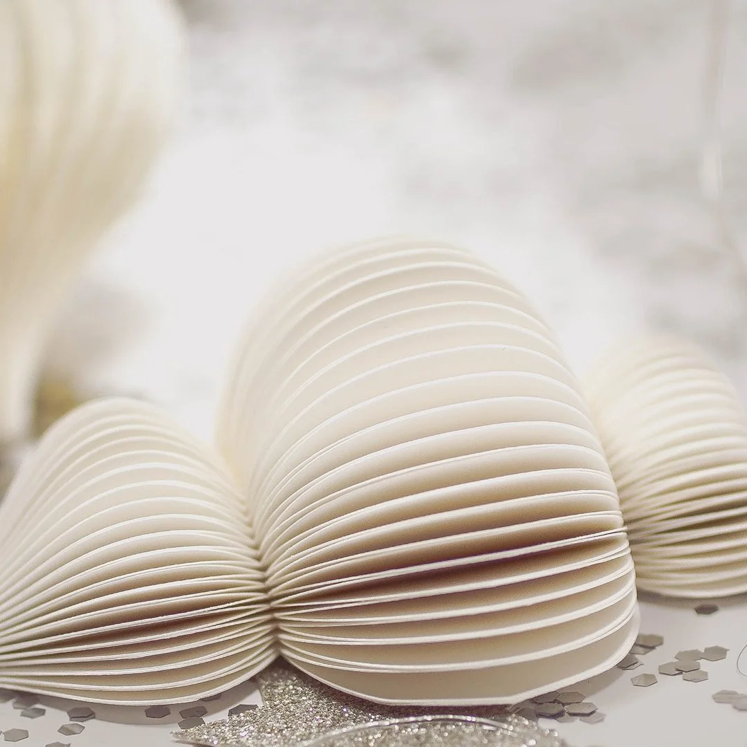 A close view of cream folded paper ornaments on a pale surface, the concertina folds catching soft light