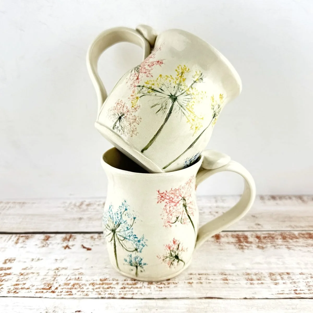 Two cream mugs stacked on a whitewashed board, pressed and painted with Queen Anne's lace in pink, blue, yellow and green