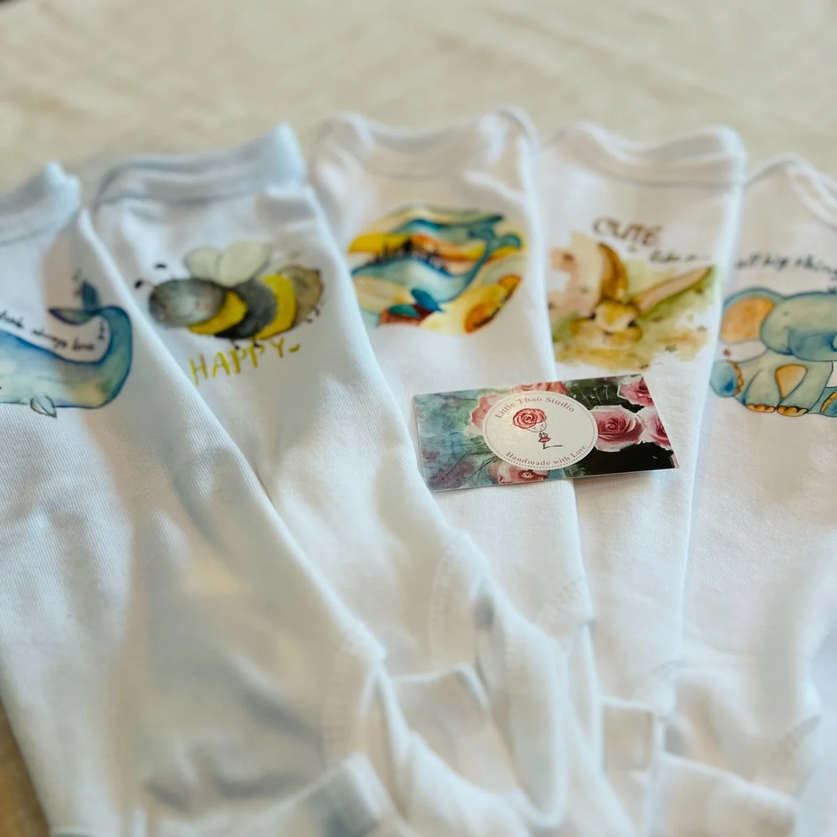 A row of white baby onesies printed with watercolour animals, a whale, a bee, a rabbit and an elephant, beside a Little Thao Studio card