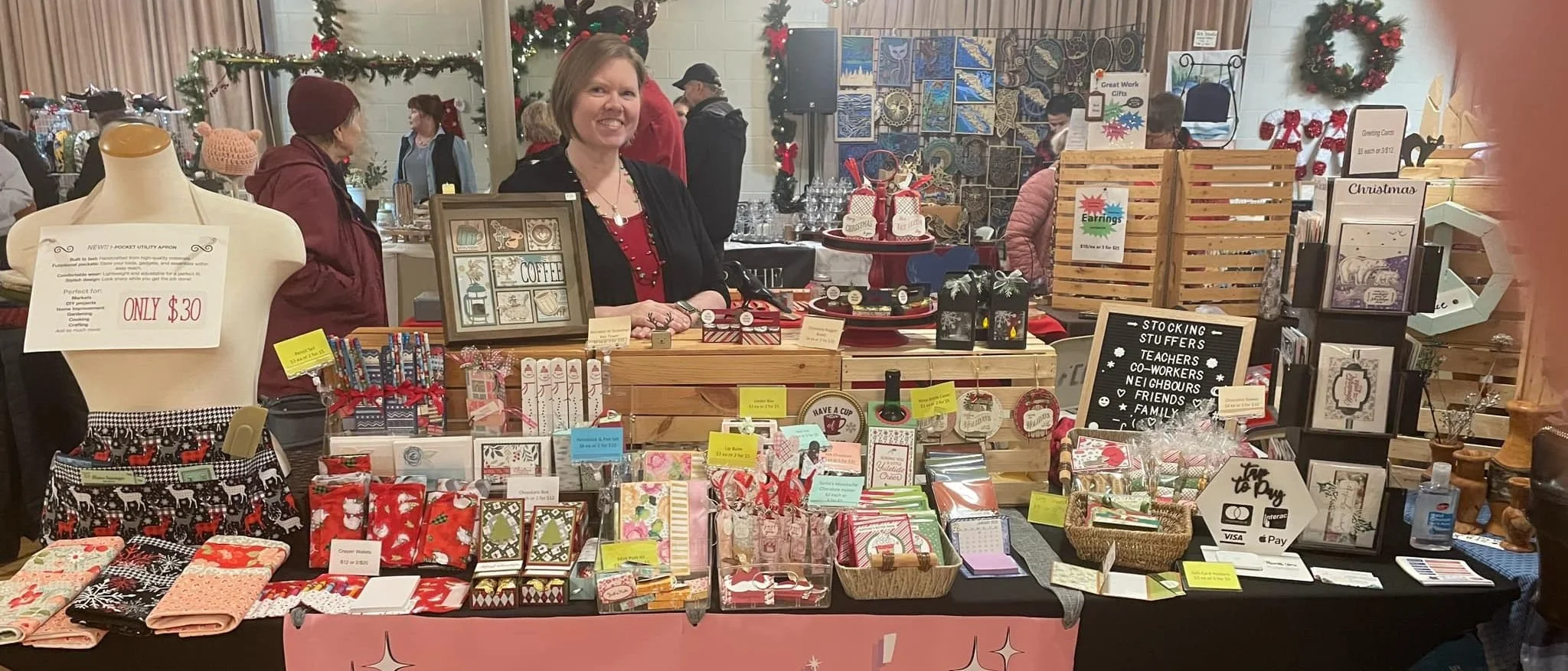 The Stampin' Queens booth at a Christmas market, a long table of cards, treat holders and small gifts, with an apron on a dress form at one end and a letter board reading stocking stuffers for teachers, co-workers, neighbours, friends and family