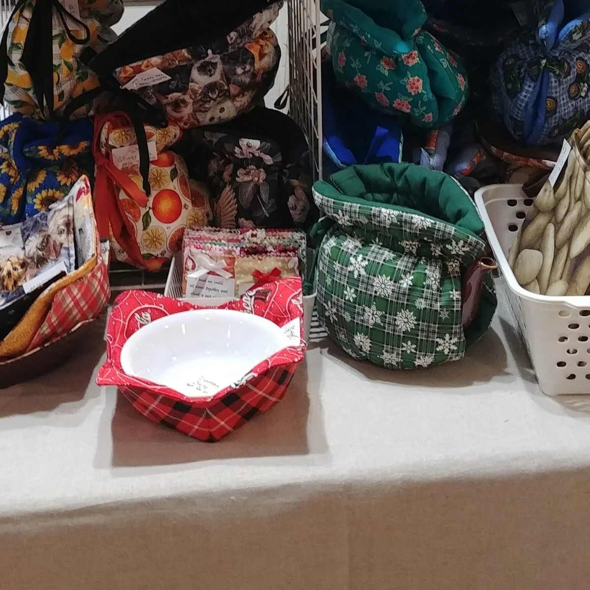 A red plaid bowl cozy holding a white bowl, beside drawstring bags in green snowflake and floral prints and a basket of quilted potato bags
