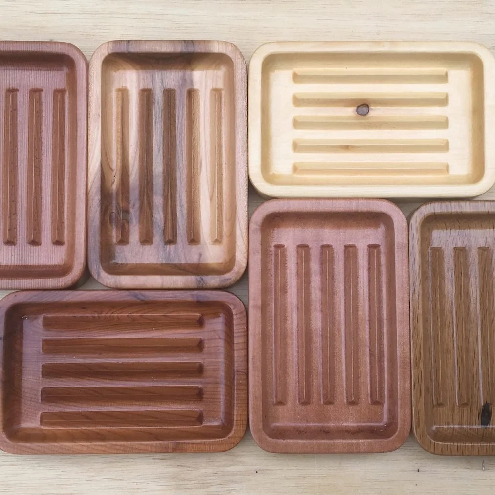 Six carved wooden soap dishes in different woods, each with drainage grooves cut into the base