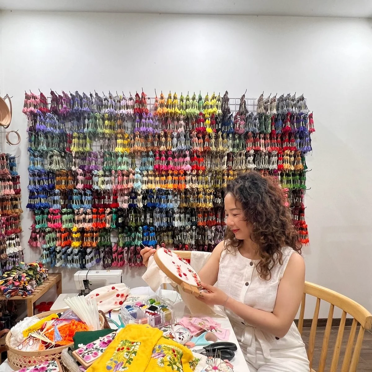 Nga stitching an embroidery hoop at her workbench in front of a wall of coloured threads