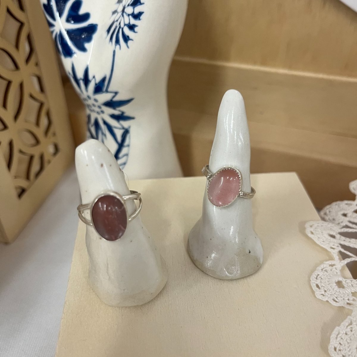 Two silver rings set with soft pink stones, displayed on small handmade ceramic finger stands beside a blue and white vase