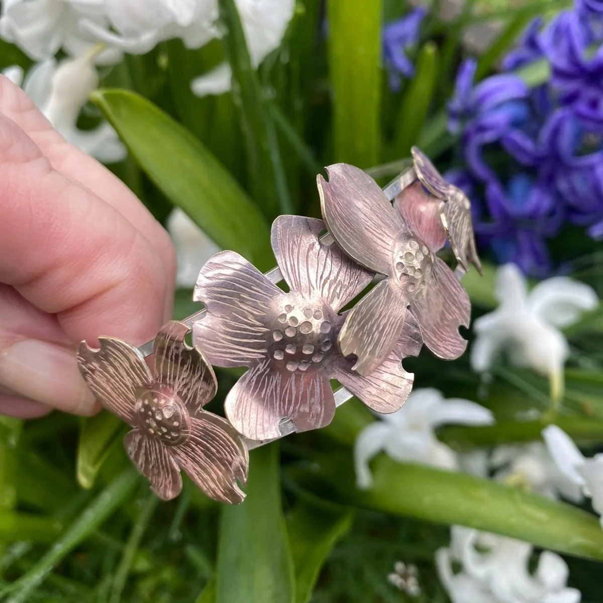 A cuff bracelet of linked dogwood flowers in copper and silver with a soft pink patina, held up in front of white and blue hyacinths