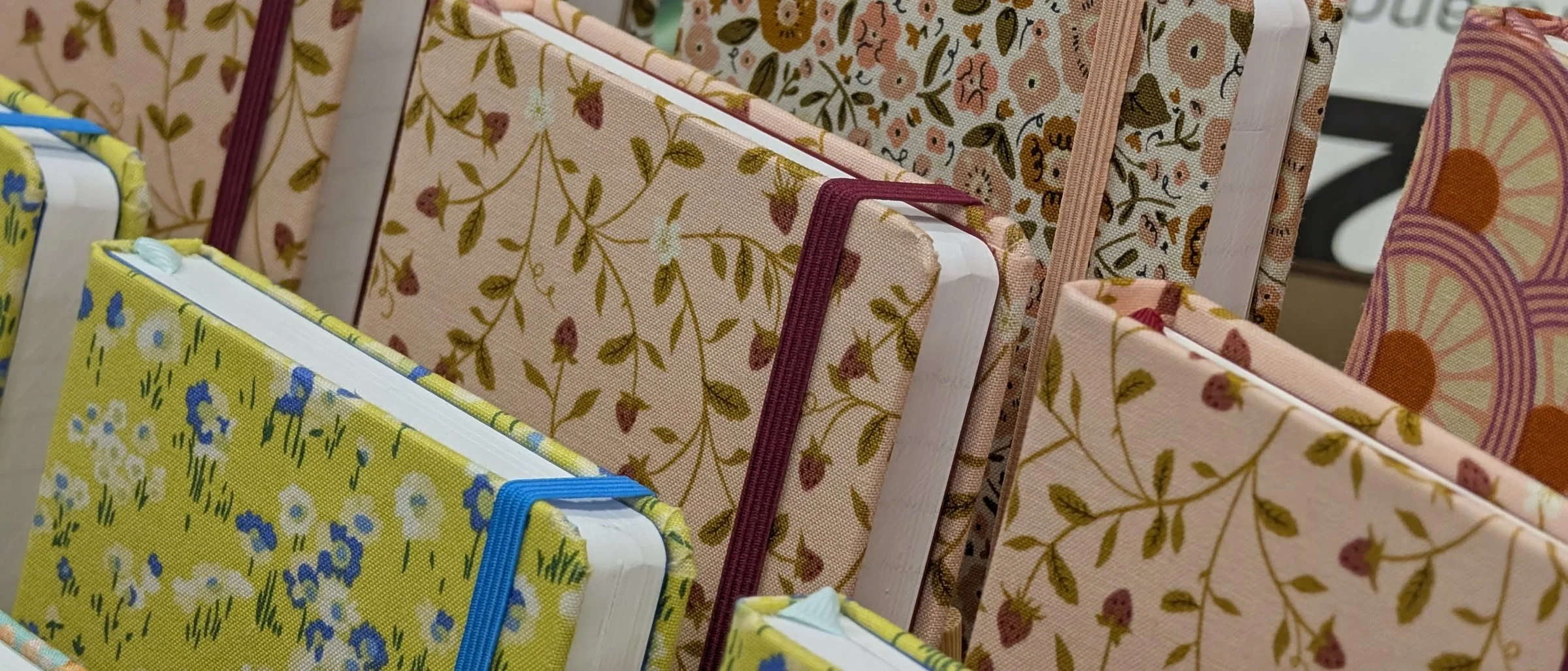 A rack of hand-bound notebooks covered in floral and patterned fabric in pinks, greens and cream, each held shut with a coloured elastic