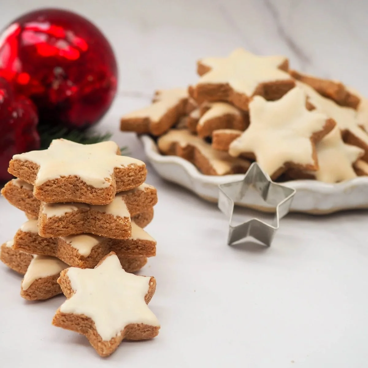 Iced cinnamon stars stacked beside a dish of more, with red glass baubles and a star cutter alongside