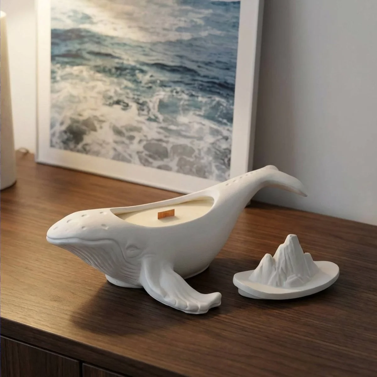 A candle in a white humpback whale vessel with a wooden wick, its lid sculpted as a small iceberg and set down beside it