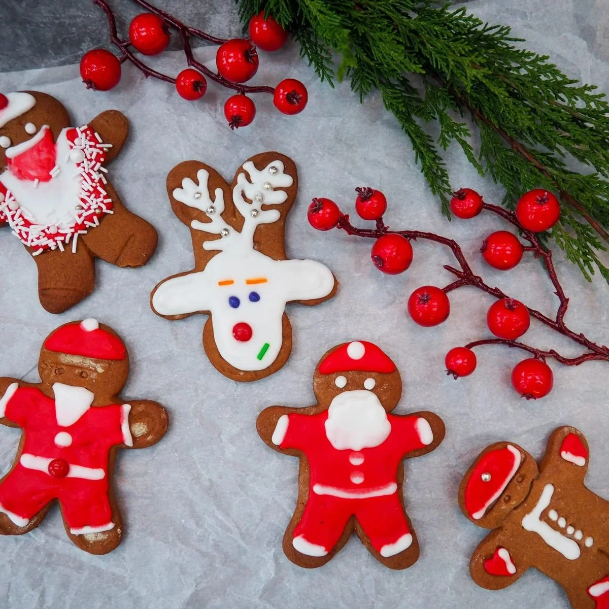 Iced gingerbread figures, a Santa, a reindeer and a snowman, laid out on parchment with red berries and cedar
