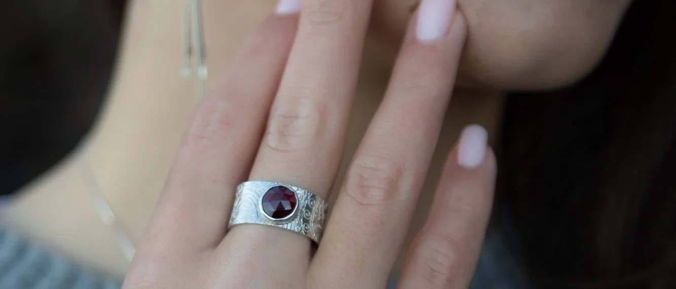 A wide sterling silver band ring set with a faceted deep red garnet, worn on a hand resting against the wearer&rsquo;s cheek, a silver and amber earring soft in the background