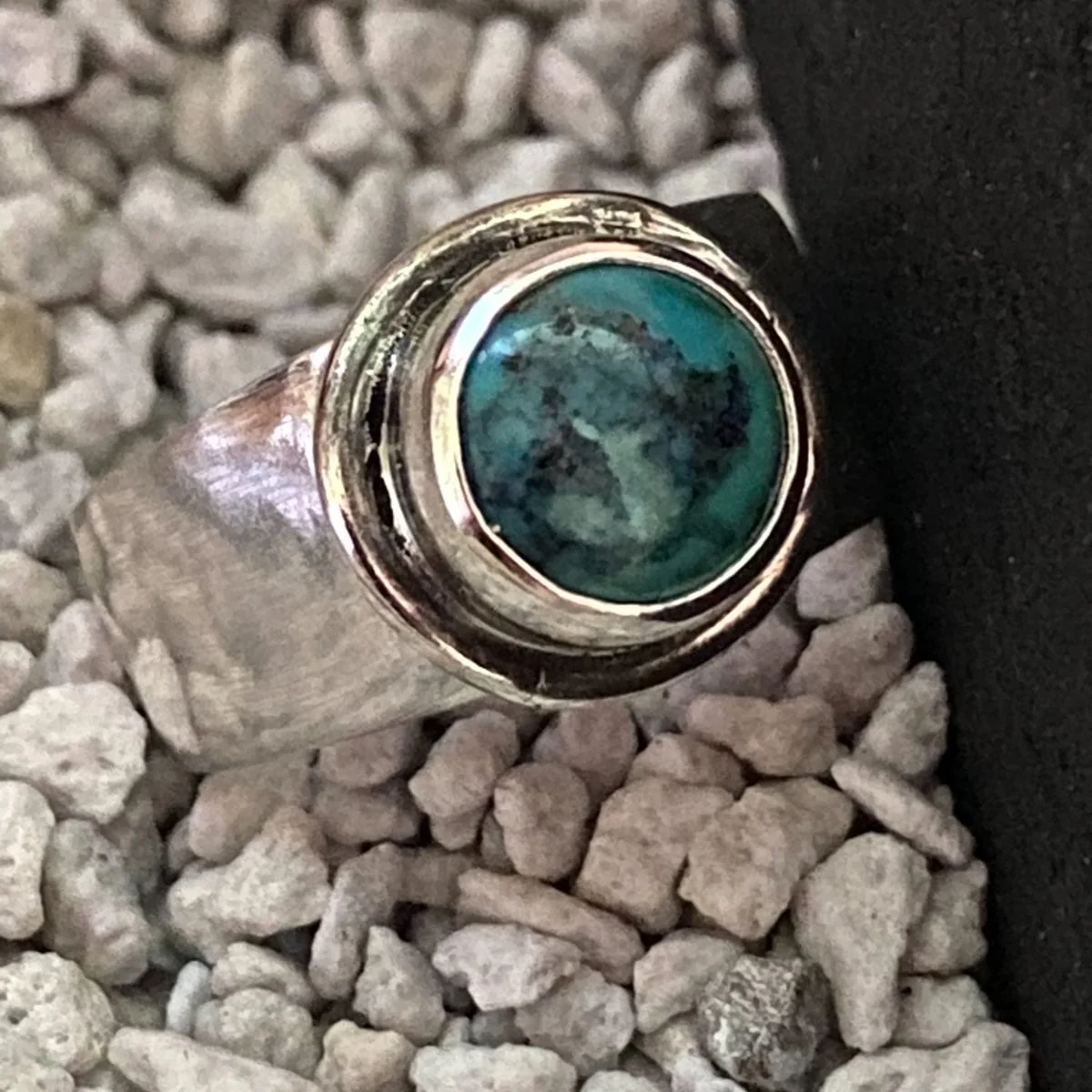 A silver ring set with a round turquoise stone flecked with dark green and black, standing on pale gravel