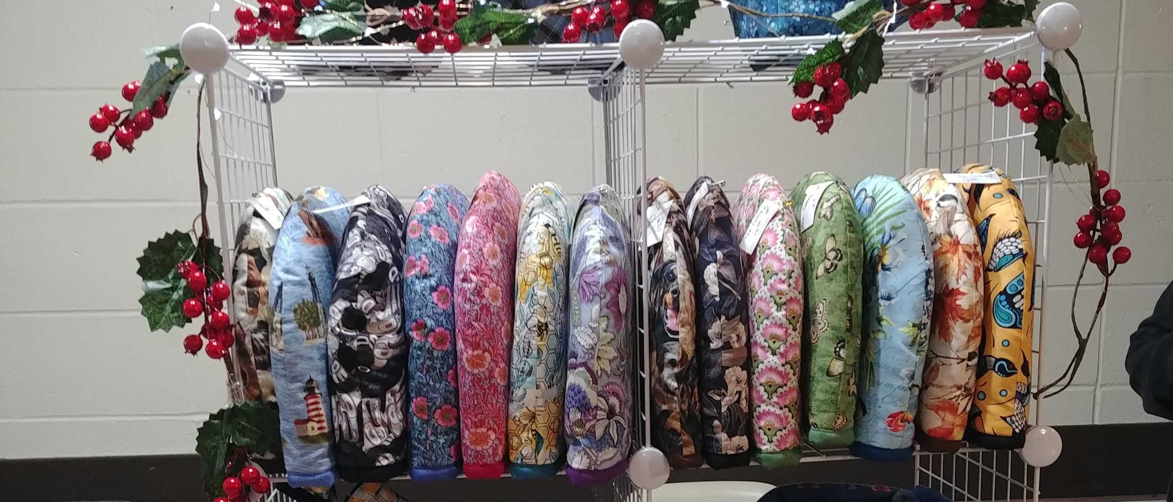 A row of quilted casserole handle covers standing on a wire shelf in florals, plaids and animal prints, framed by a garland of red winter berries
